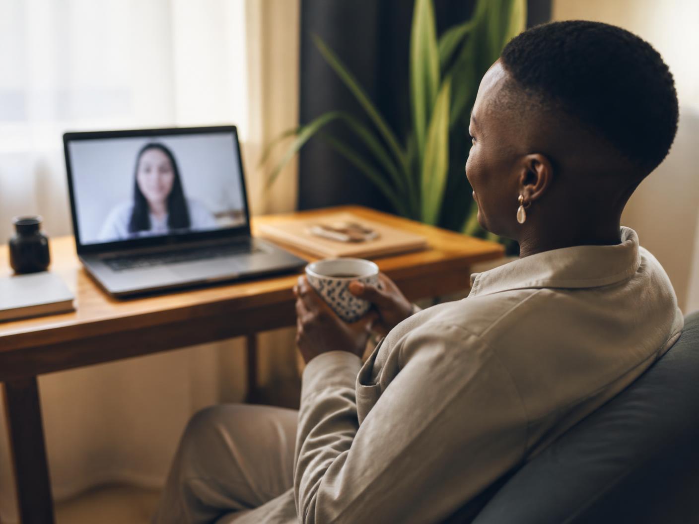 An adult seated comfortably at home, holding a warm drink while speaking with a mental-health professional over a laptop video call